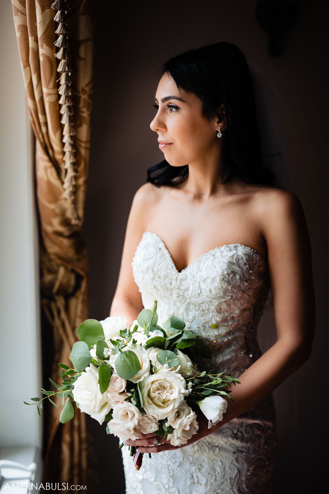 Elegant bride in lace strapless gown holds white and green bouquet while standing by window with gold curtains.