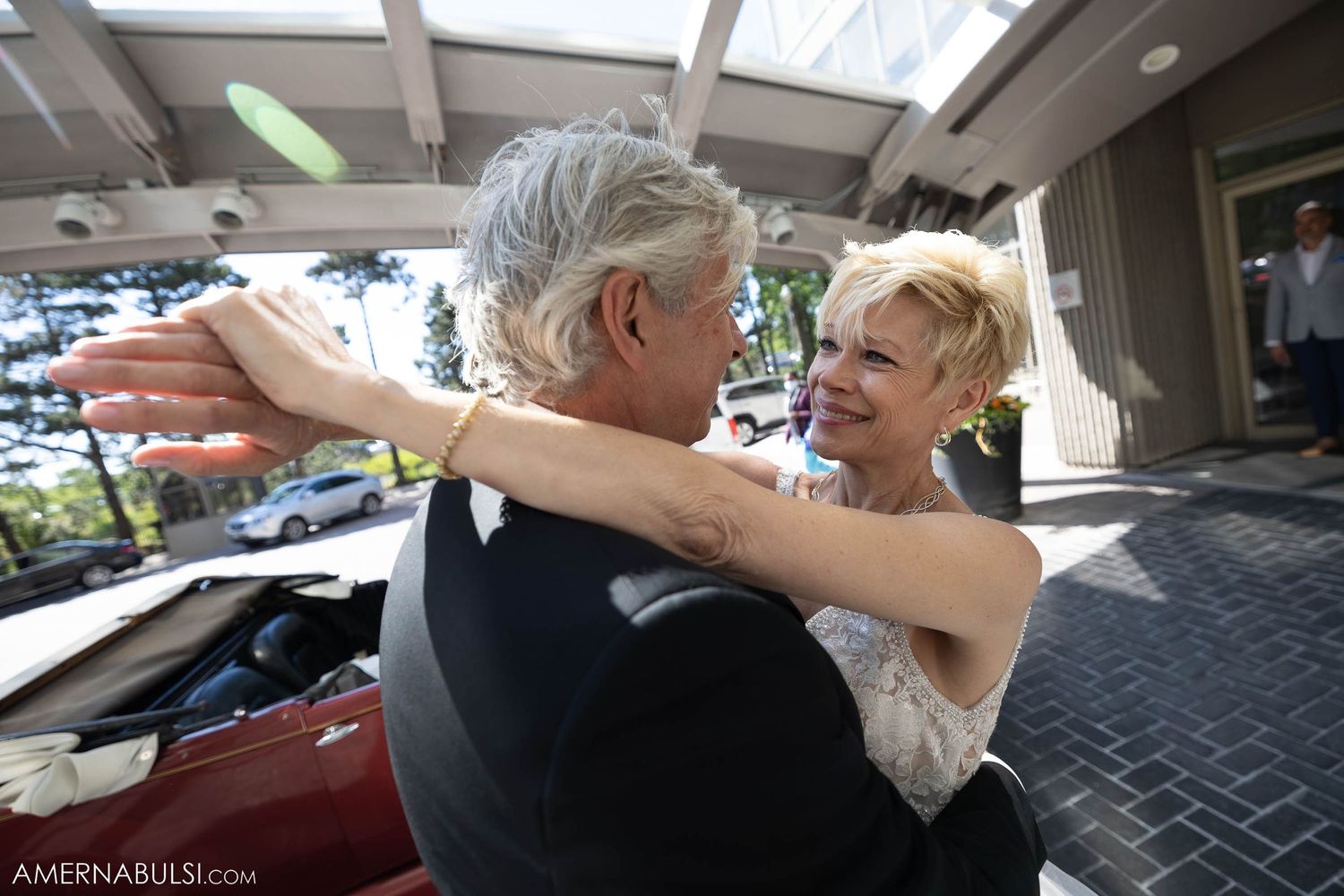 A couple embracing in formal attire next to vintage automobiles.