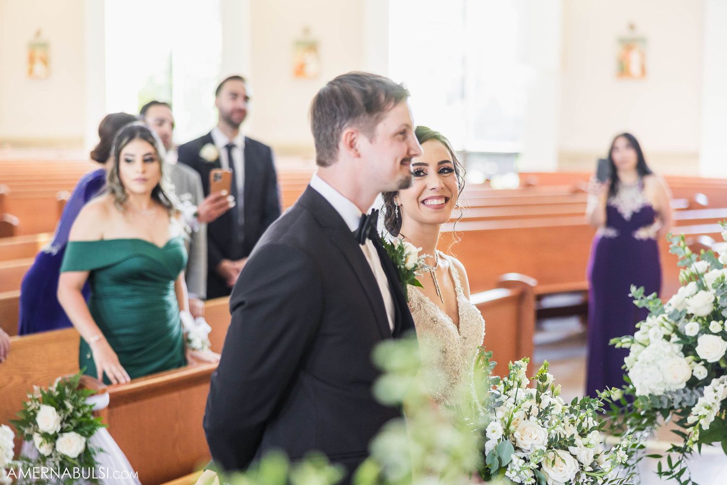 Wedding party members in formal attire gather inside an elegant church with white floral arrangements.