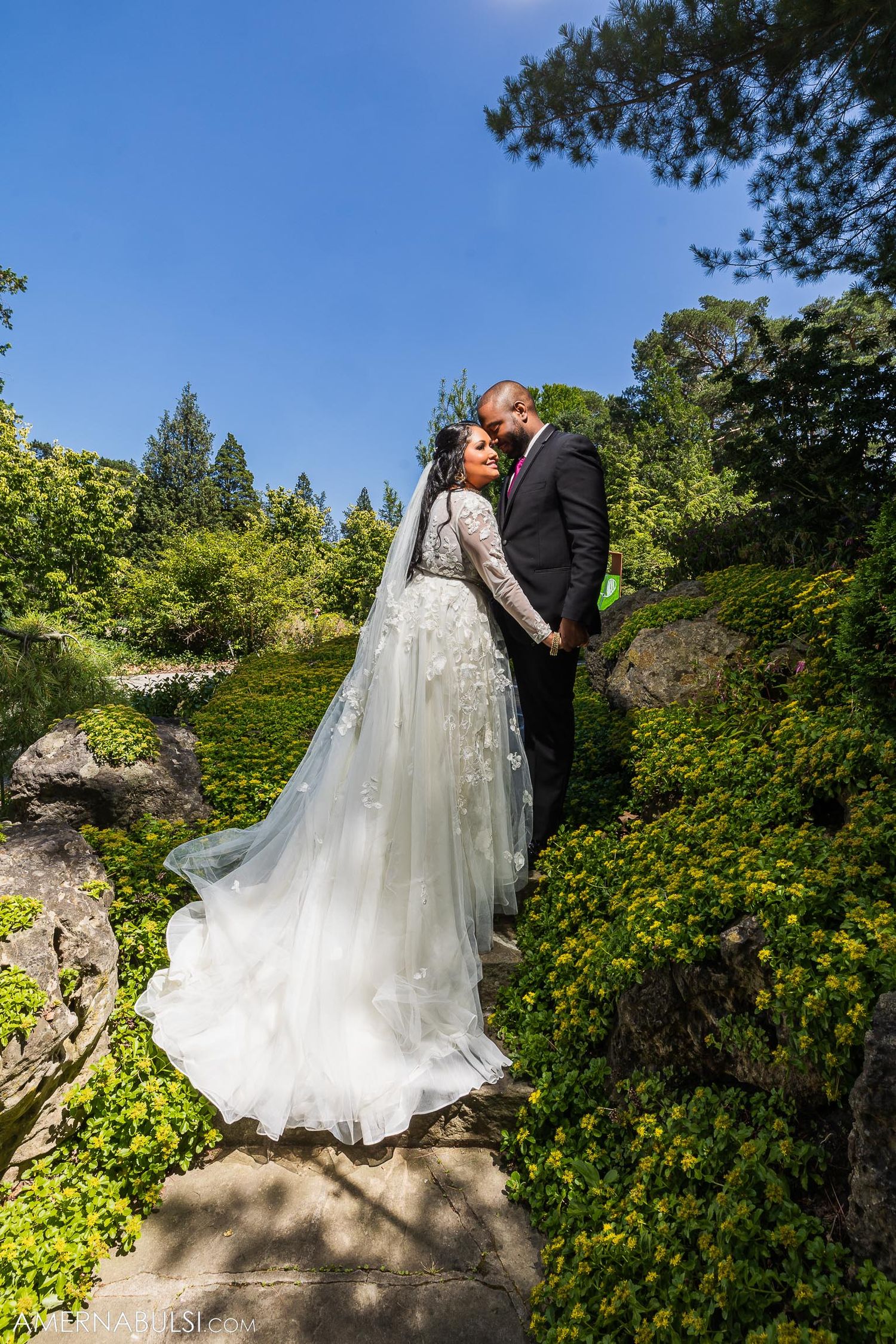 Couple in wedding attire standing on garden path among yellow flowers and greenery.