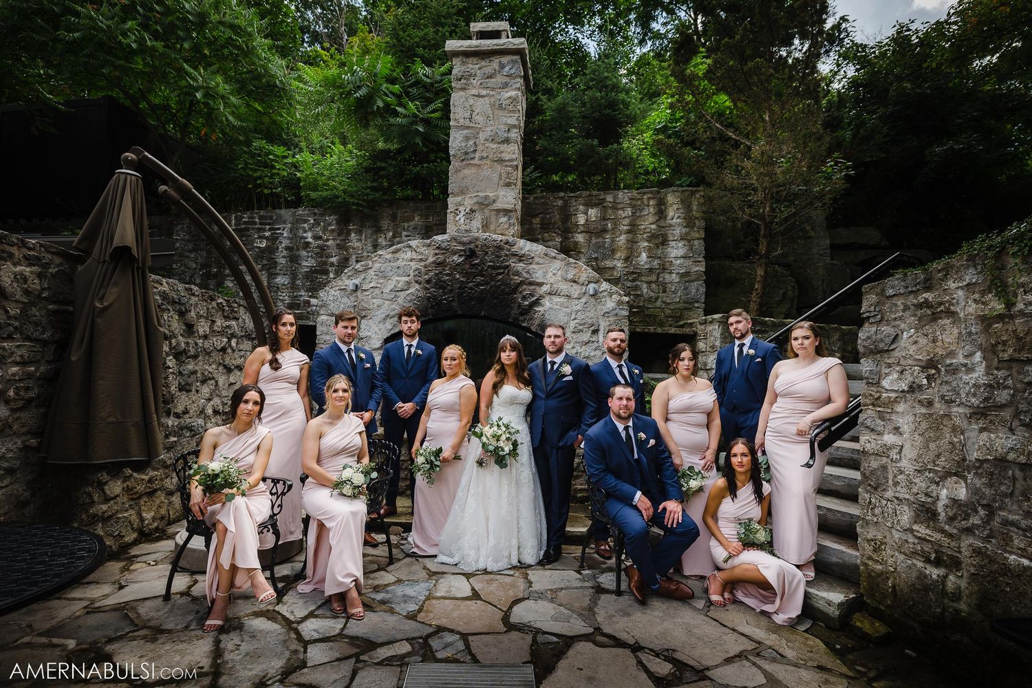 Wedding party poses on stone steps against rustic wall backdrop in formal attire.