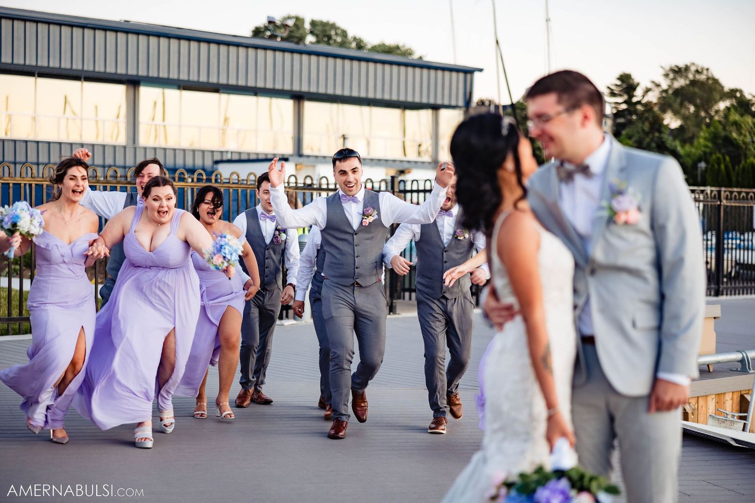 Wedding party walks along marina dock with bridesmaids in purple dresses and groomsmen in grey suits.