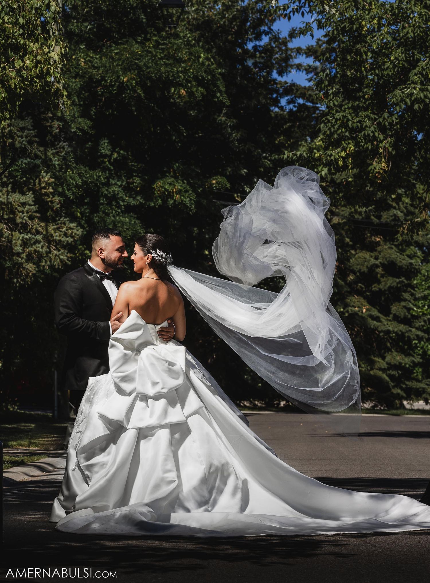 A bride's veil flows dramatically in the wind as the couple embraces outdoors.
