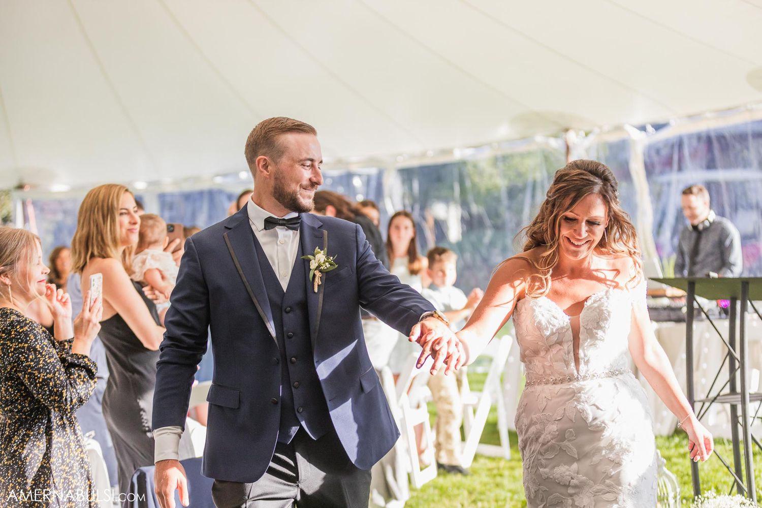 Joyful newlyweds exit their wedding ceremony under a tent while guests celebrate with smiles and applause.
