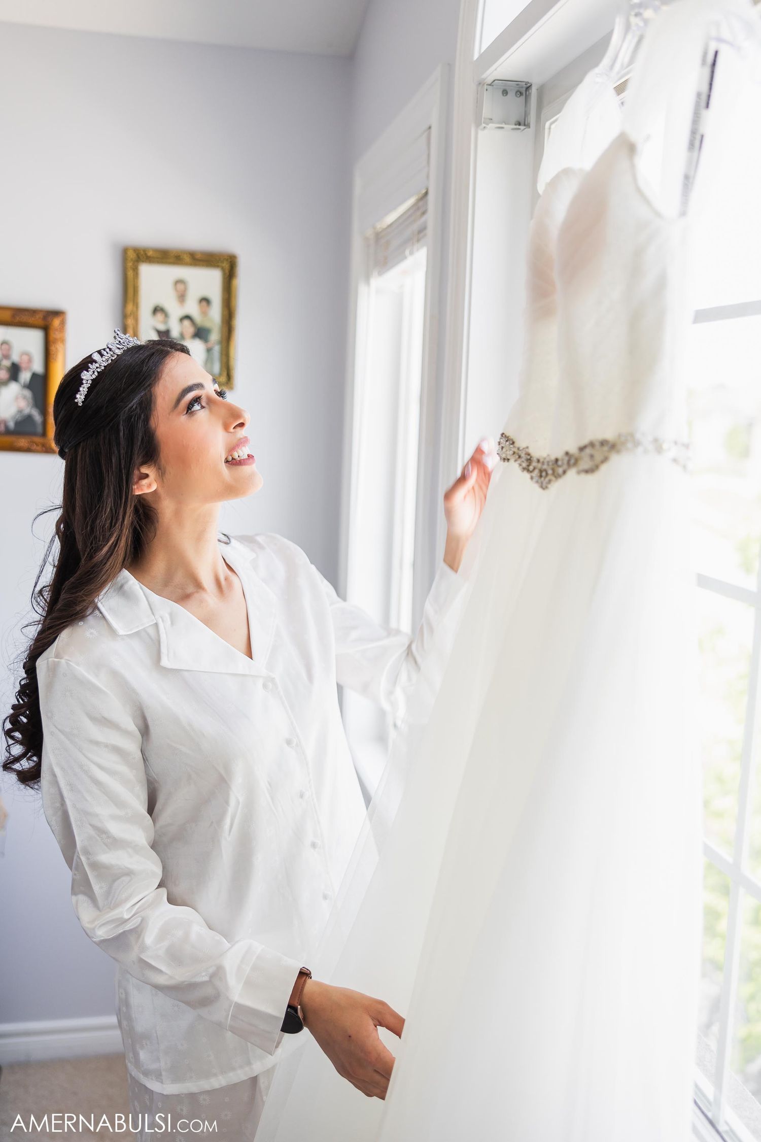 Person in white robe admires wedding dress hanging by window in morning light.