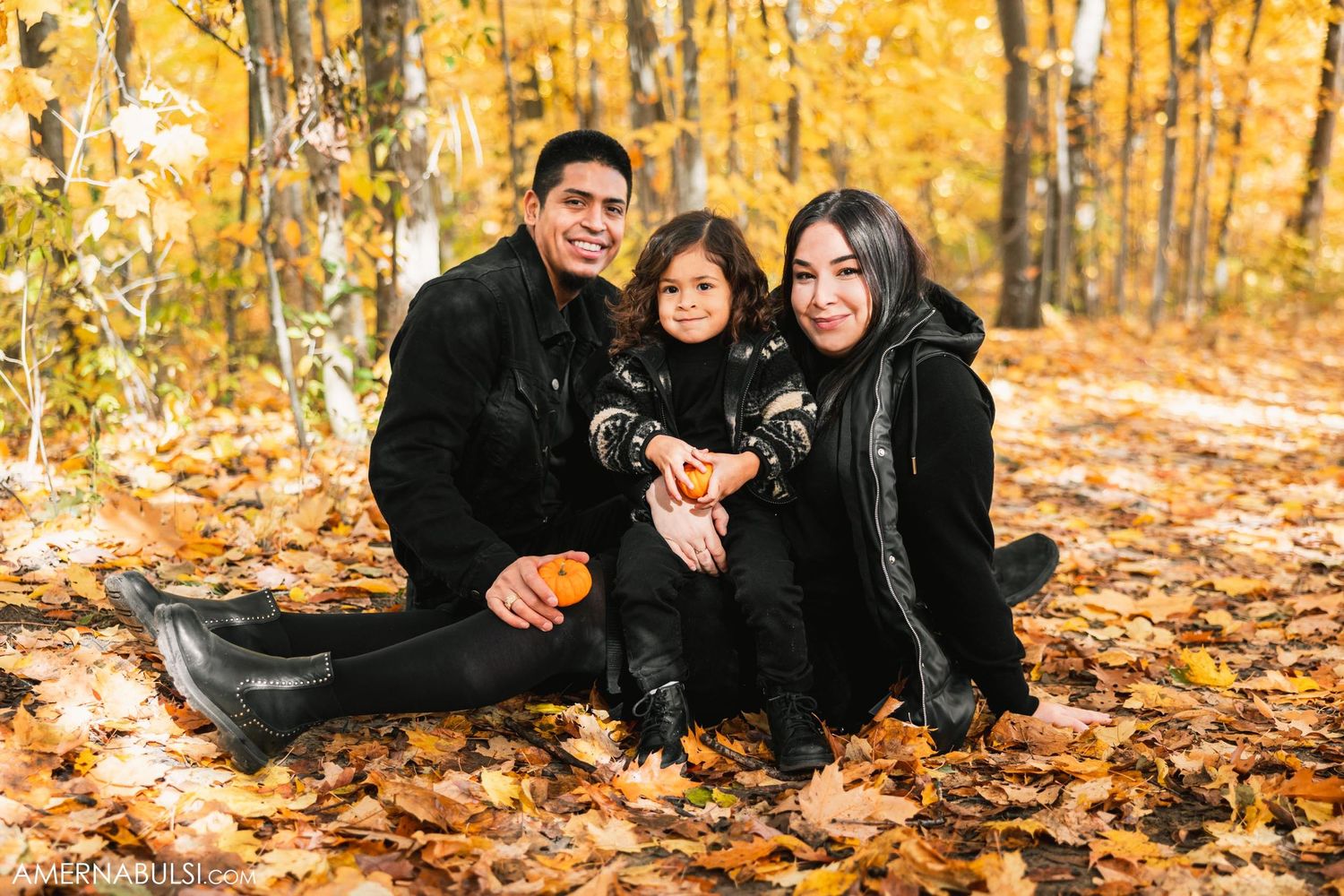 A family group sits together on fallen autumn leaves in a forest with golden foliage.