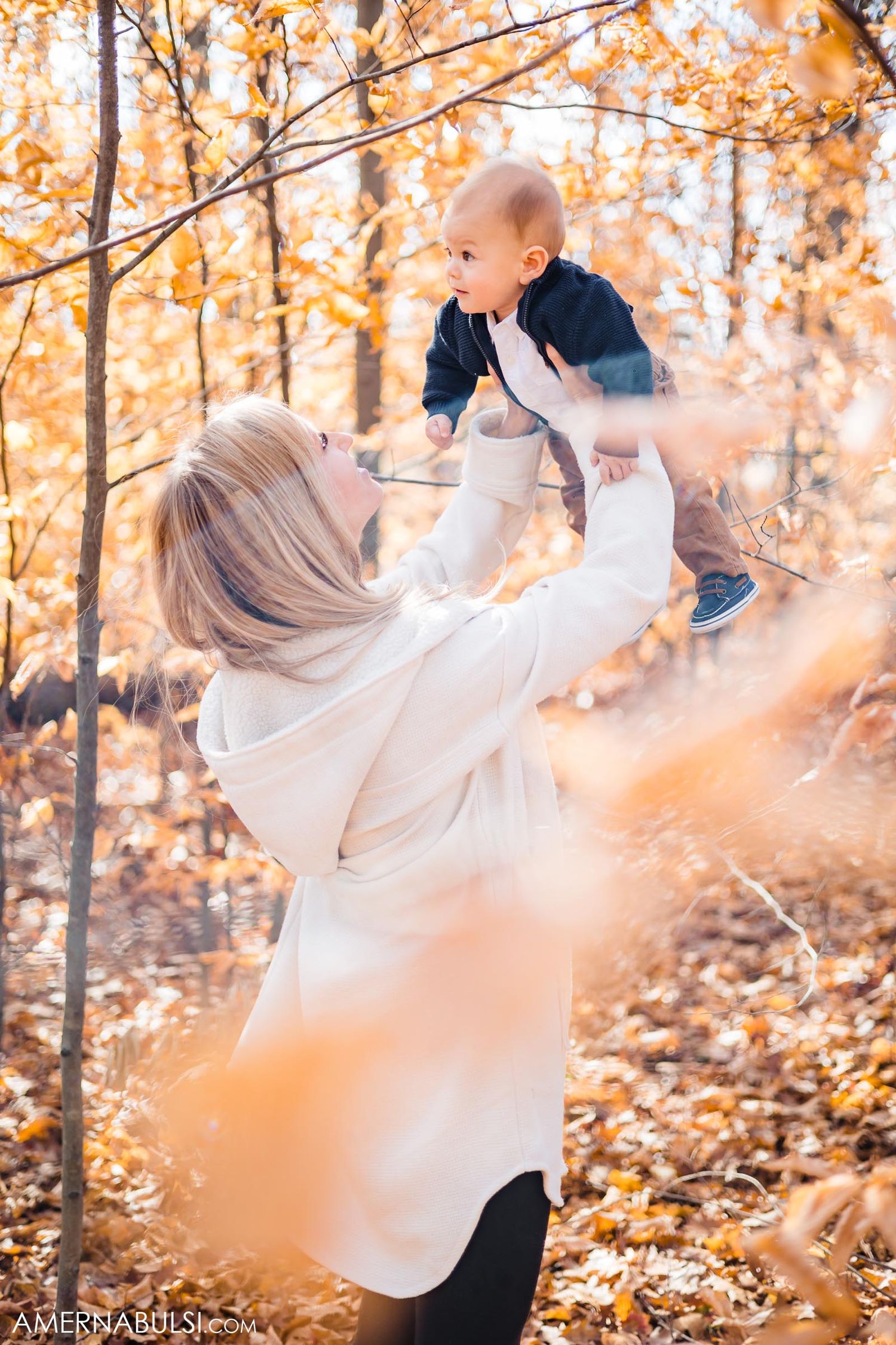 Mother lifts smiling baby high in air among golden fall foliage.