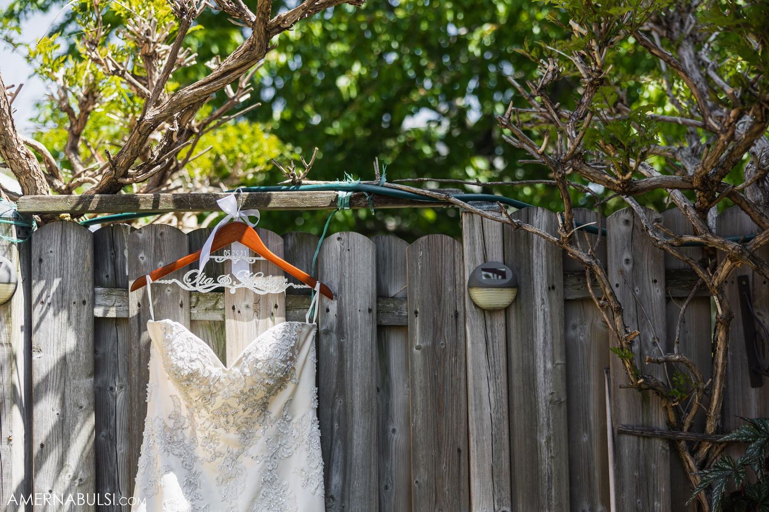 White wedding dress hanging on a rustic wooden fence with green foliage in the background.