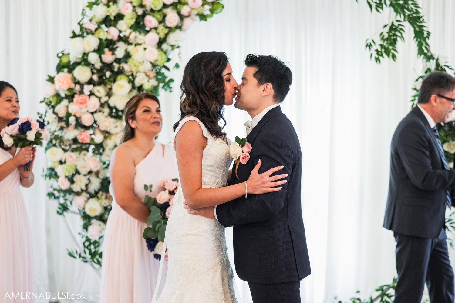 A wedding ceremony takes place under an elegant floral arch with blush and white roses.