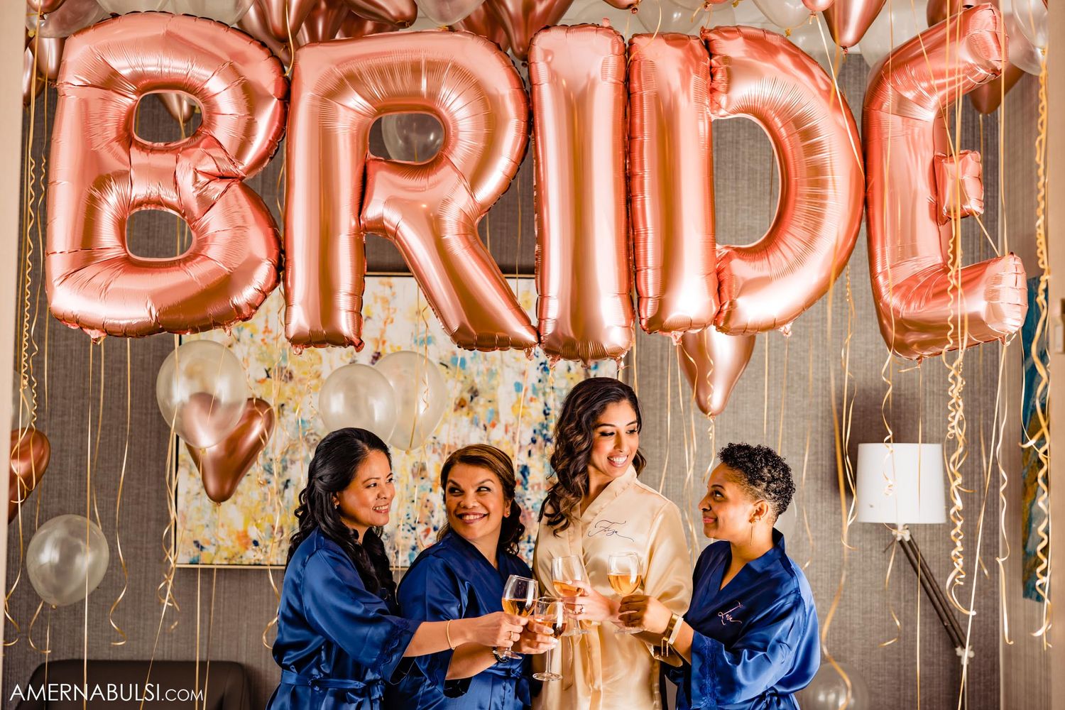 Celebratory moment with bridal party in blue robes beneath rose gold balloon letters.