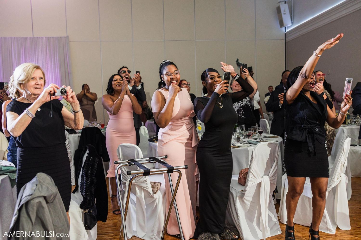 Wedding guests dancing and celebrating with raised arms at reception with decorated chairs.