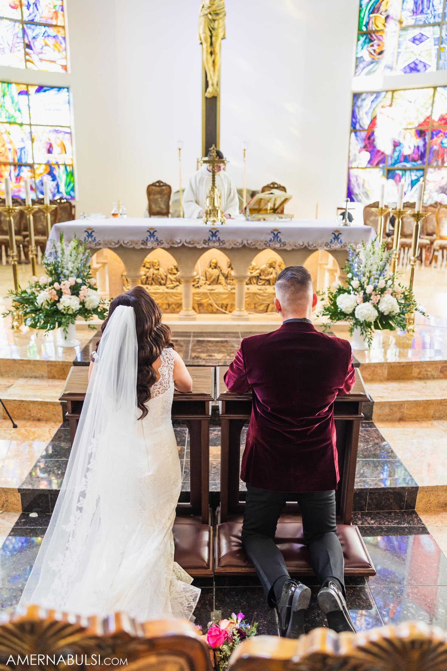 A couple kneels at an ornate church altar with stained glass windows and floral arrangements.