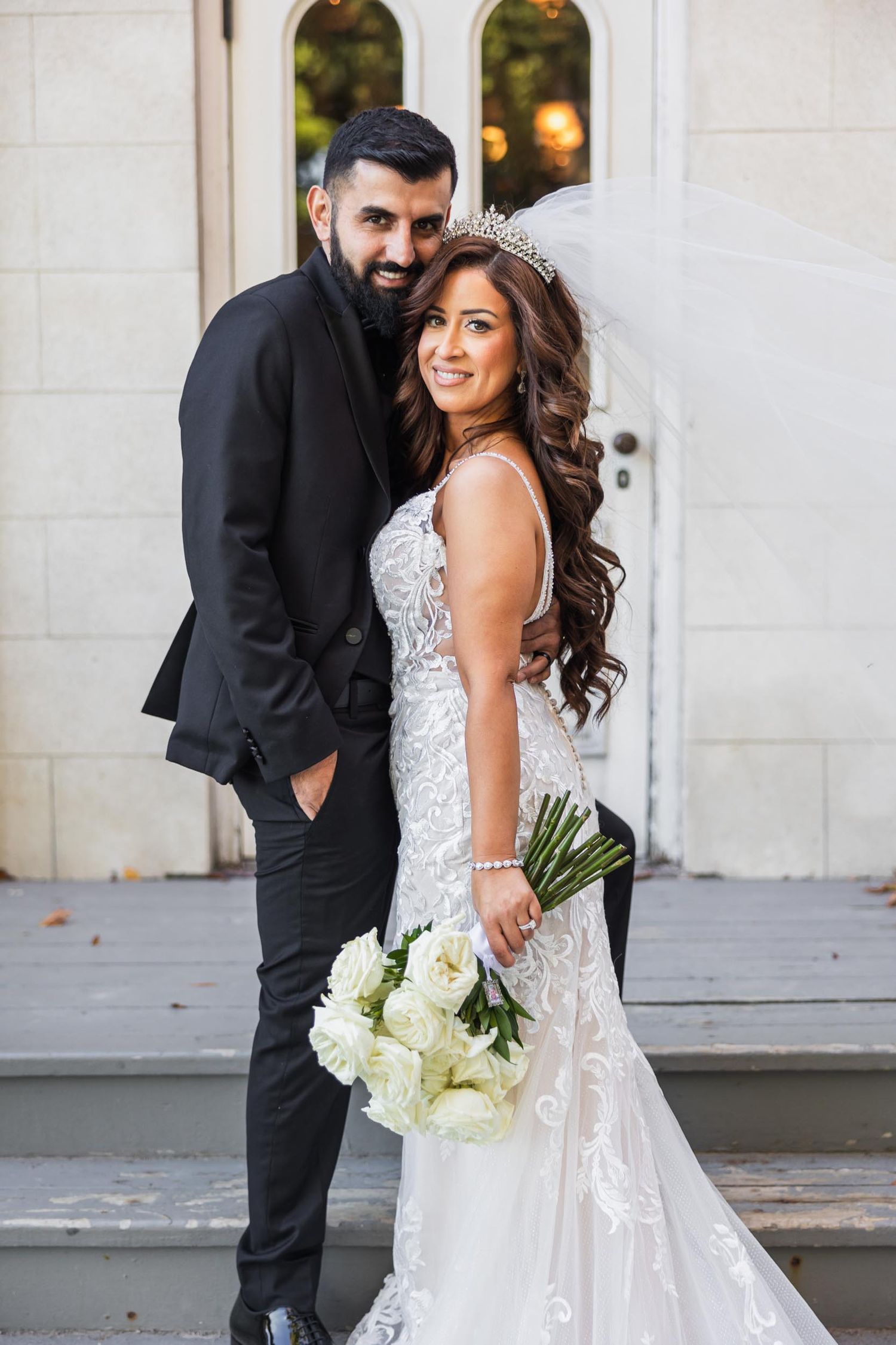 Elegant wedding couple poses against white arched doorway with bride holding white rose bouquet.