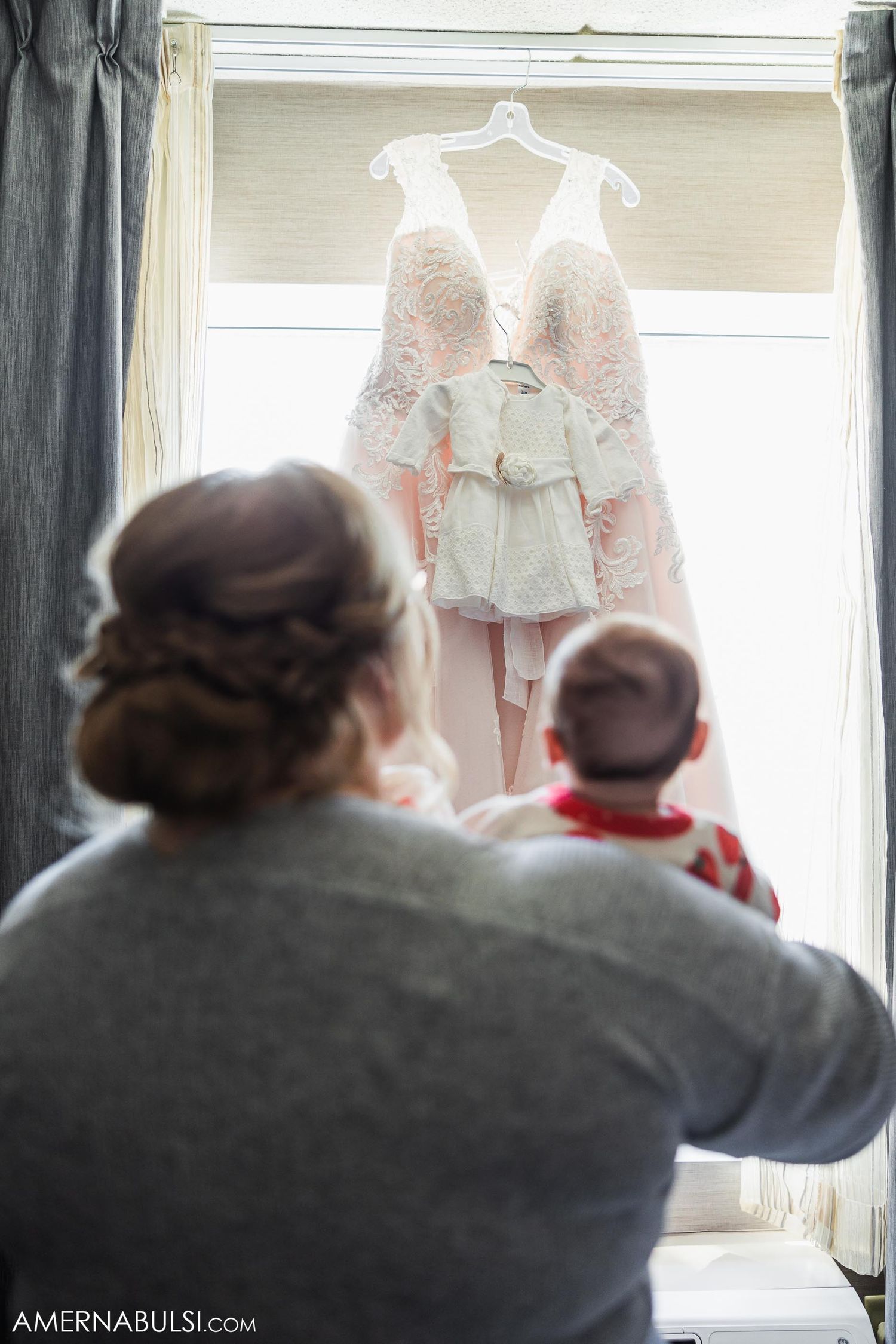Wedding dress and small white outfit hanging in window while being admired.