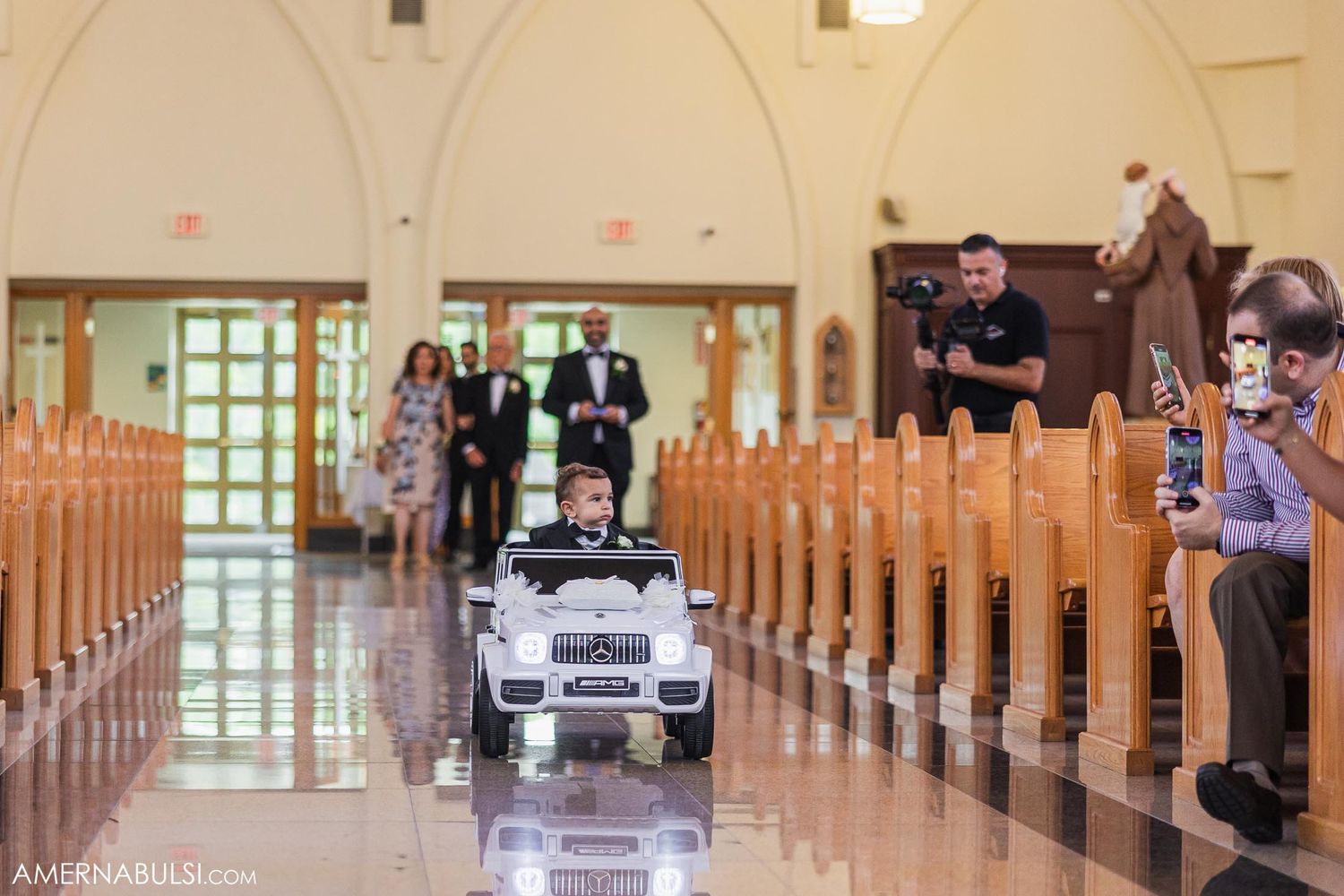 A child drives a miniature white toy car down the center aisle of a church during a wedding ceremony.