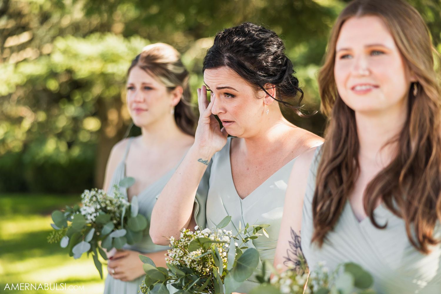 Bridesmaids in light dresses holding bouquets during an emotional outdoor wedding moment.