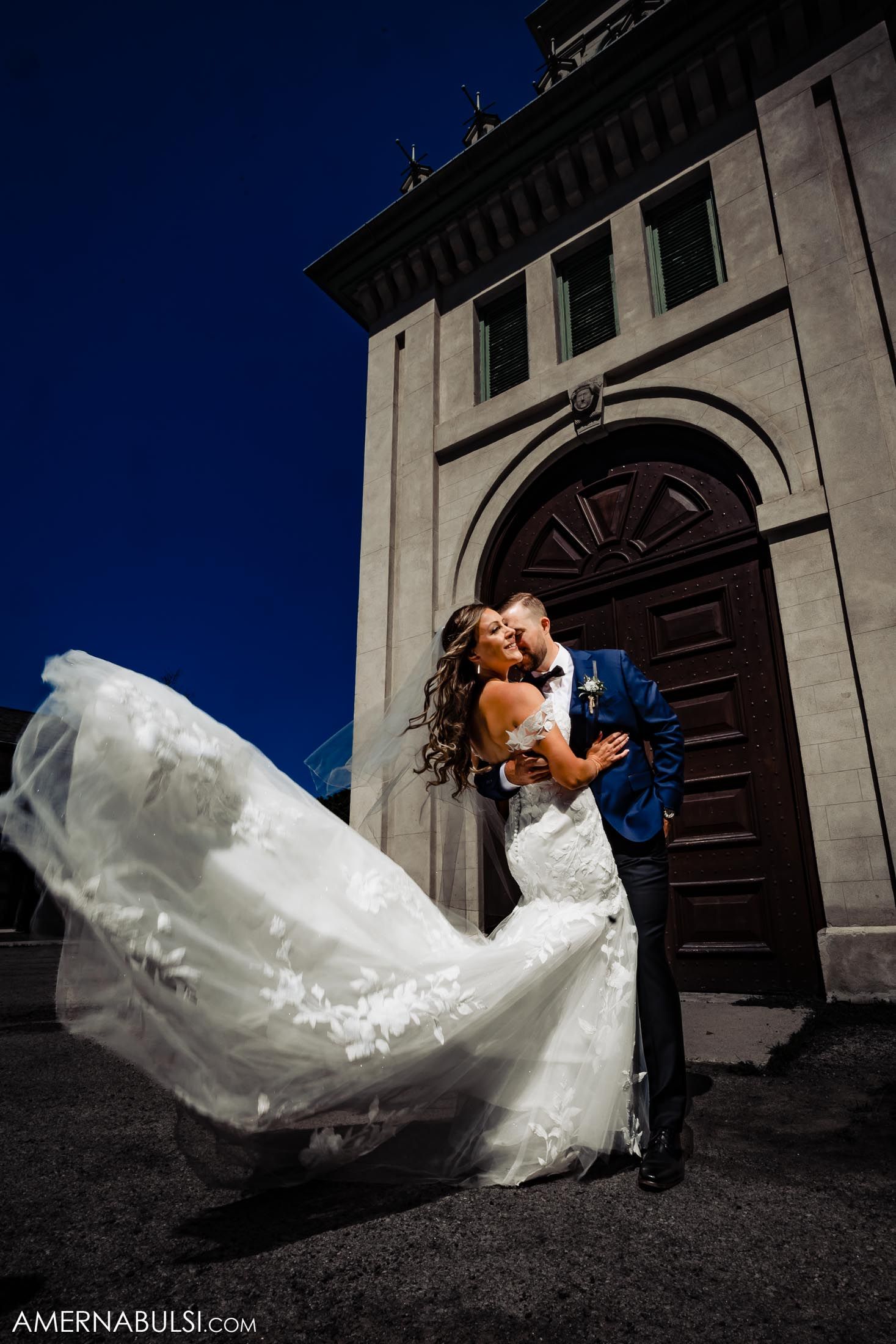 Dramatic wedding photo with flowing white dress against historic stone building entrance.