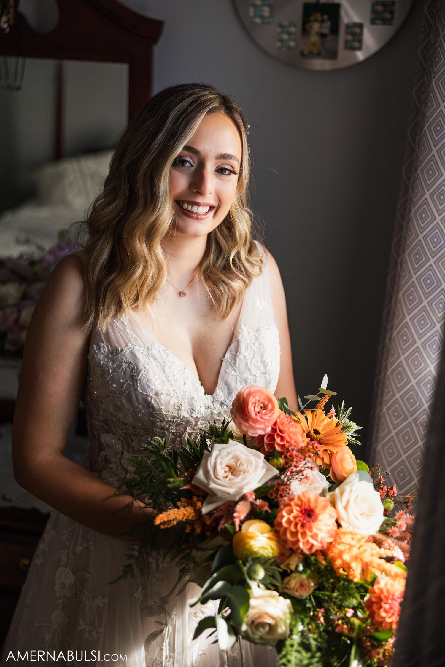 A bride poses with her coral and orange floral bouquet while standing in natural window light.