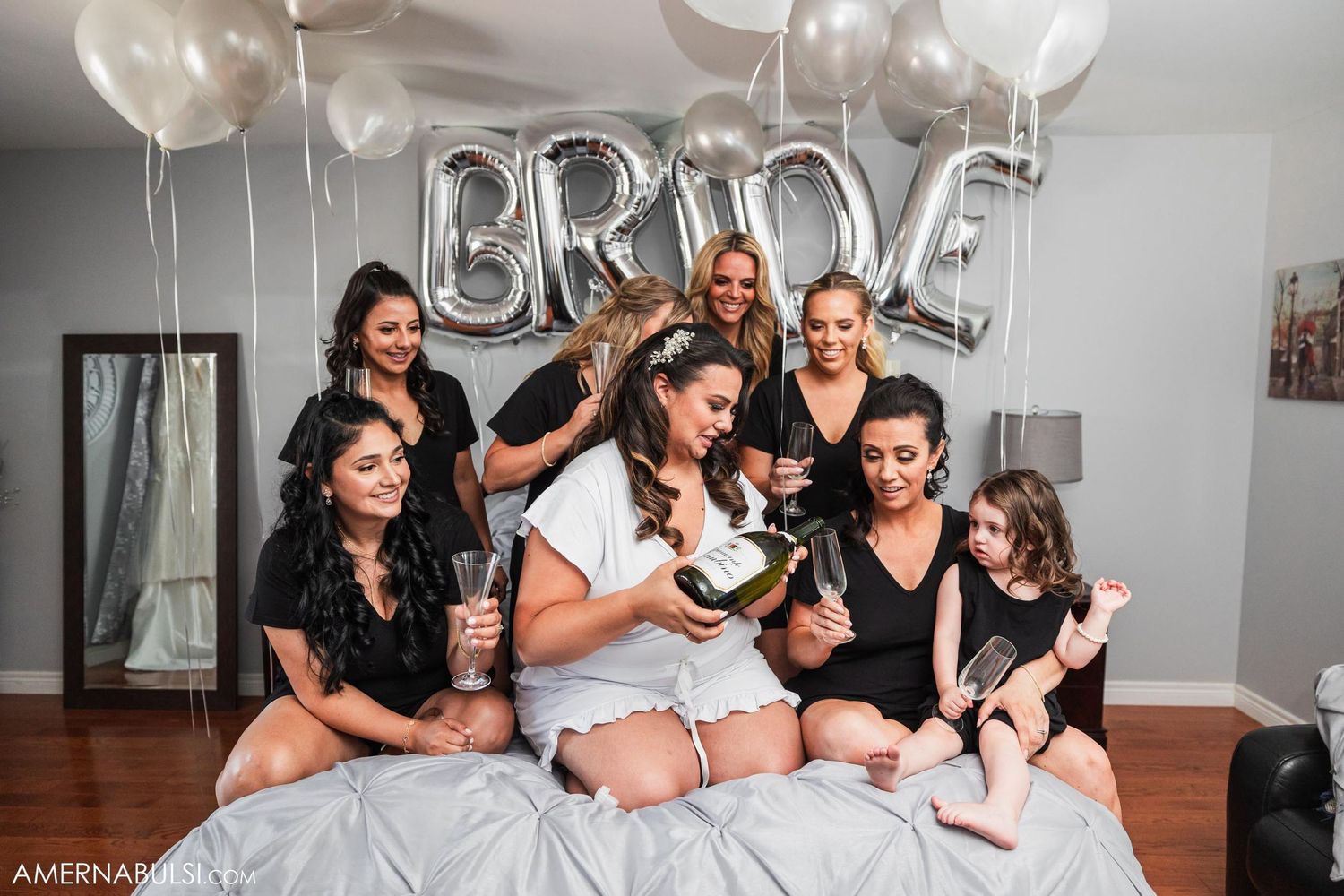 Celebrating bridal party in black robes gathered on bed with balloons and champagne.