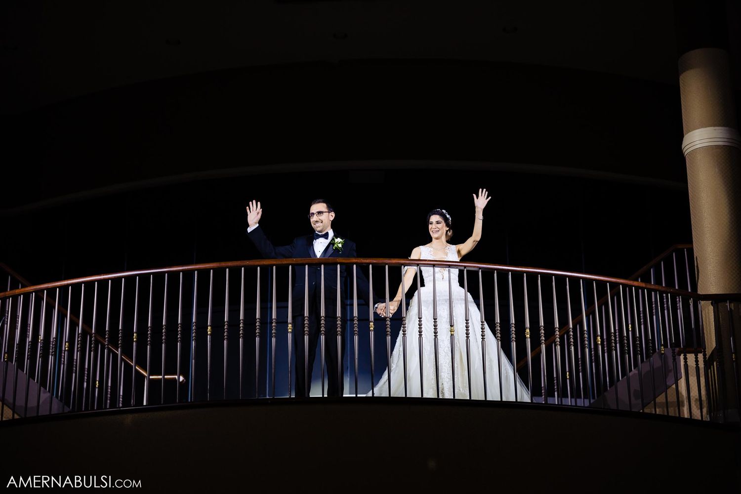 Wedding party waving from balcony railing in dramatic nighttime lighting.