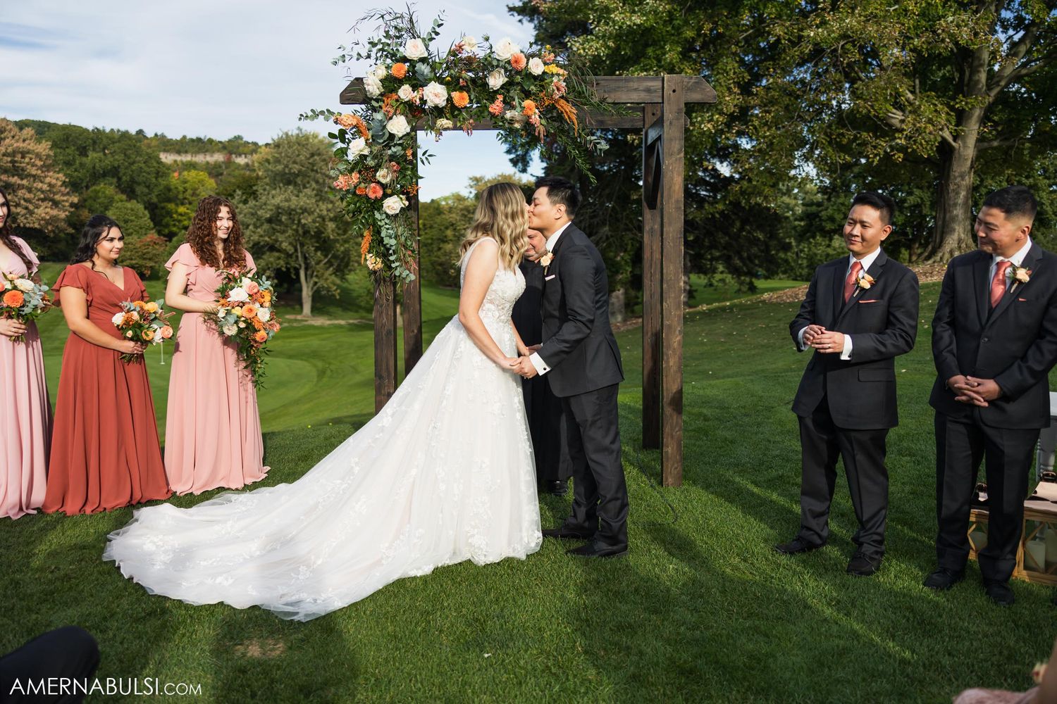 Outdoor wedding ceremony under wooden arch with pink floral arrangements.