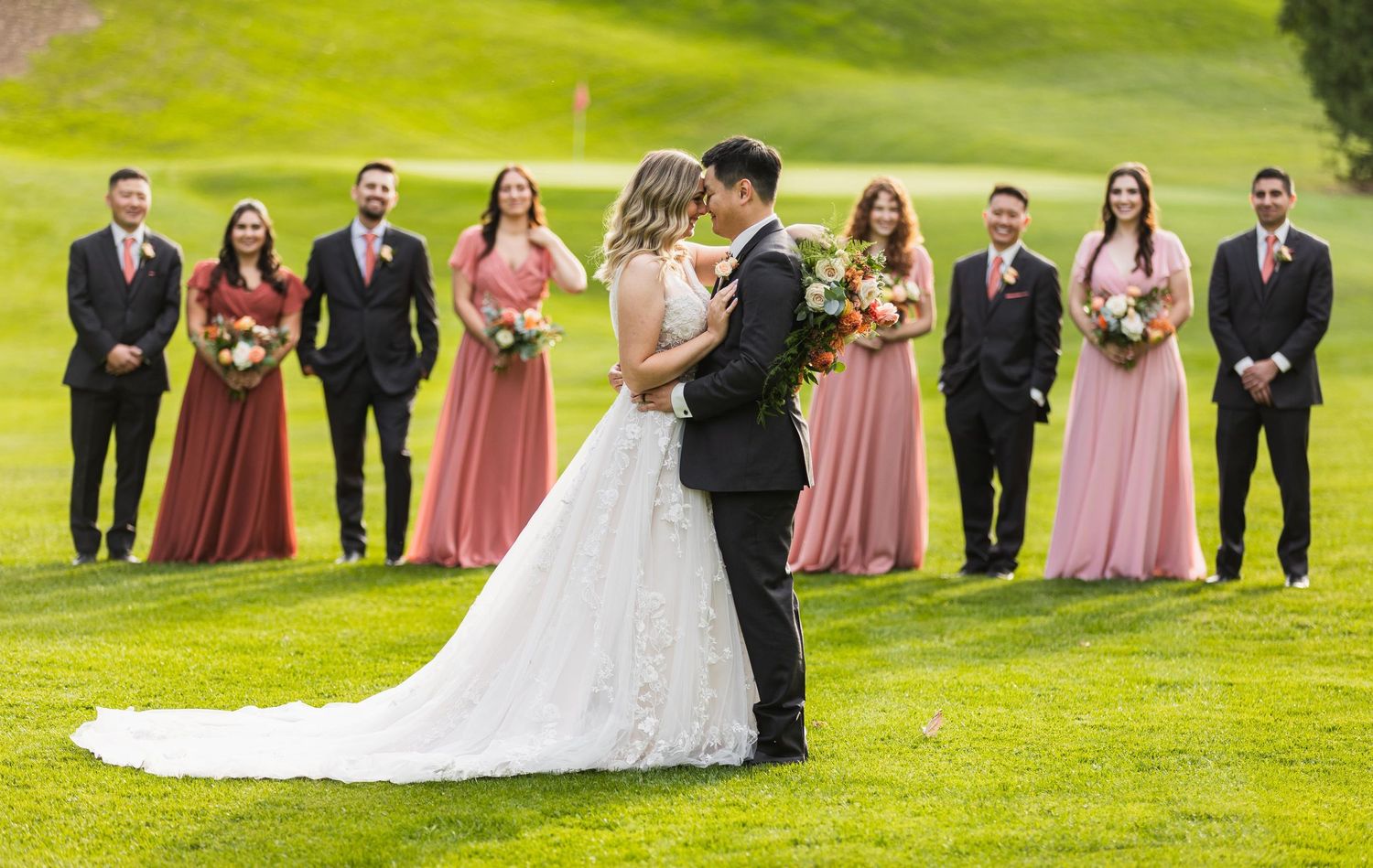 Wedding party posed on rolling green lawn with bridesmaids in pink dresses.