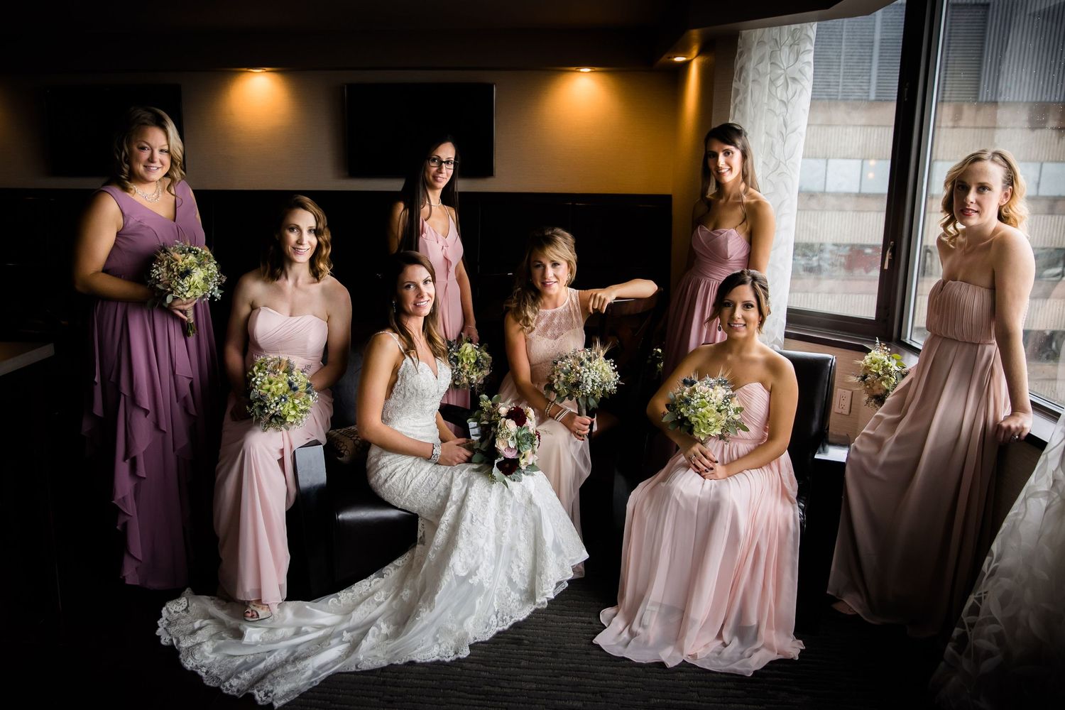 Wedding party in blush pink and plum colored dresses pose together by a window with natural light.