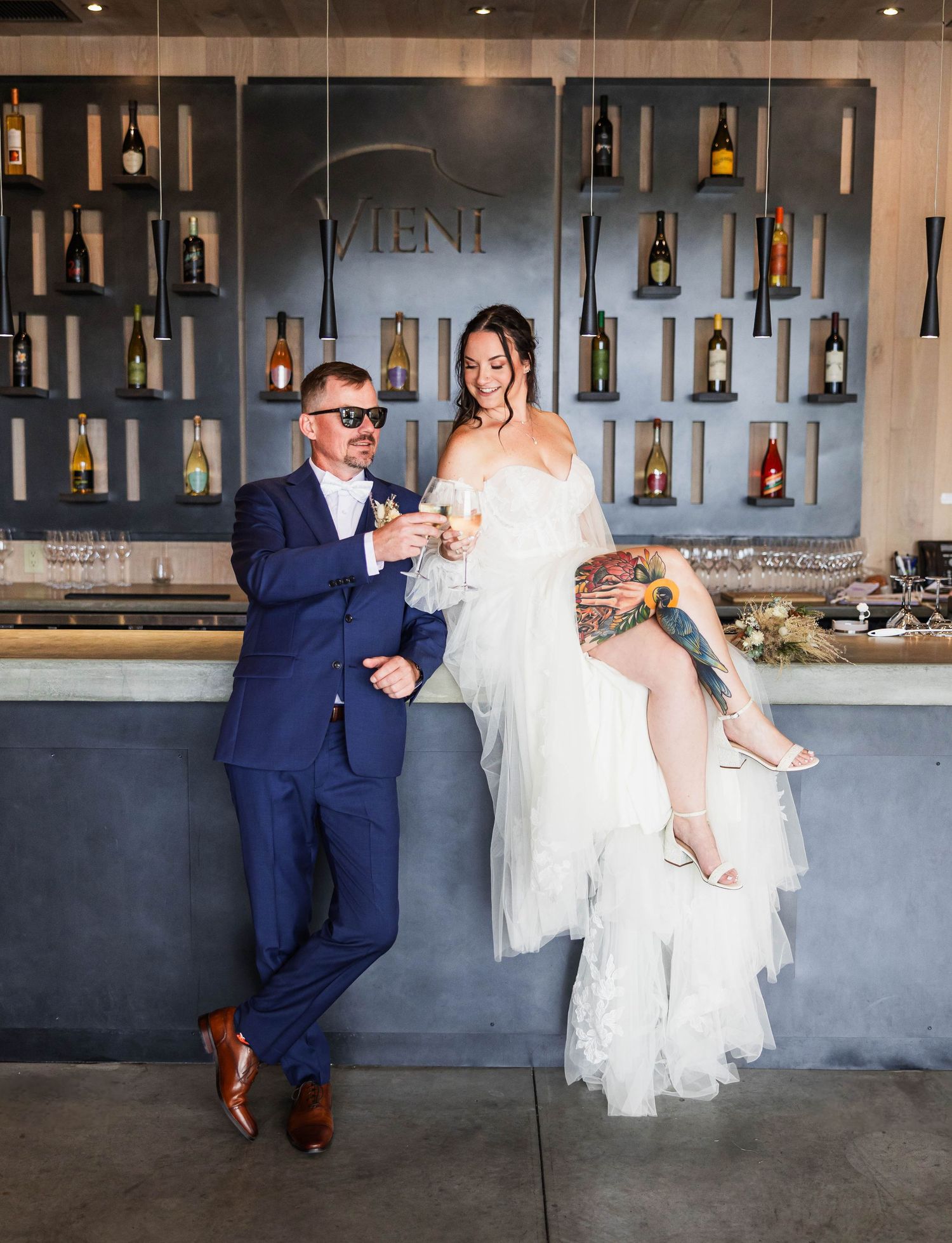 Playful wedding couple posing at modern bar with wine bottle display.