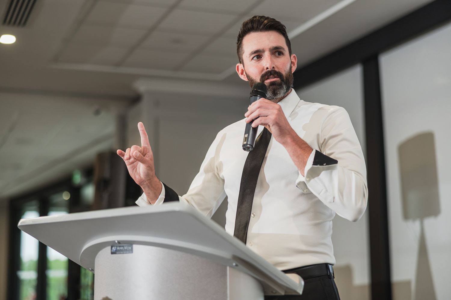 Bearded man in white shirt delivers a passionate wedding speech at a podium with a microphone.