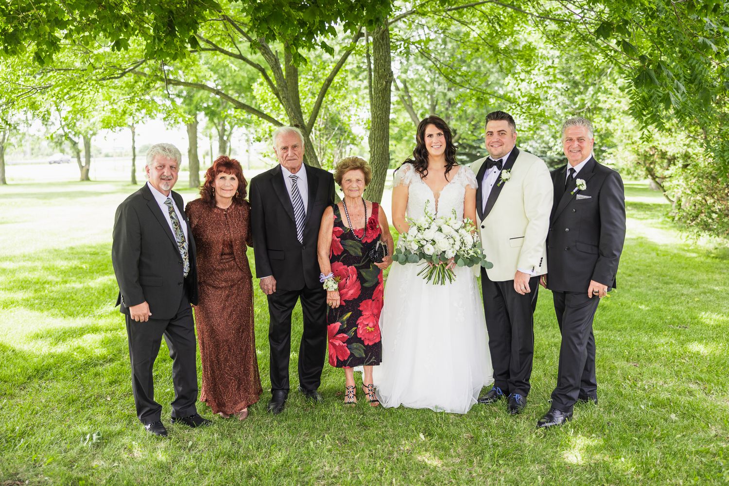 Bride and groom posing with five family members under shady trees on a sunny day.