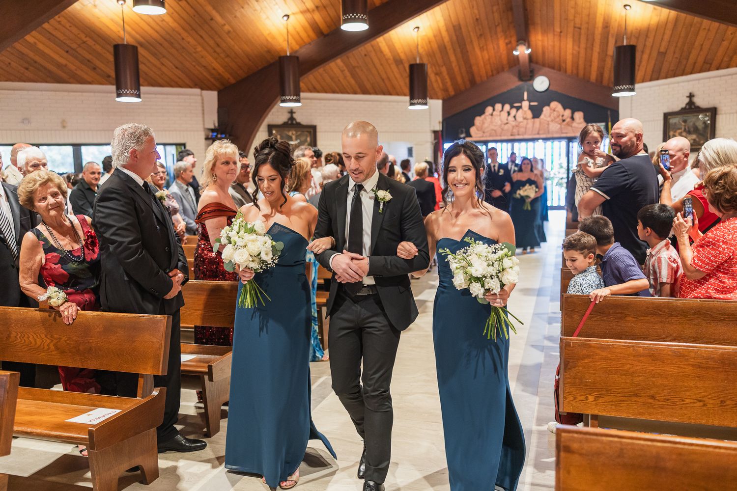Bridesmaids in teal dresses escort groomsman down church aisle during wedding ceremony procession.