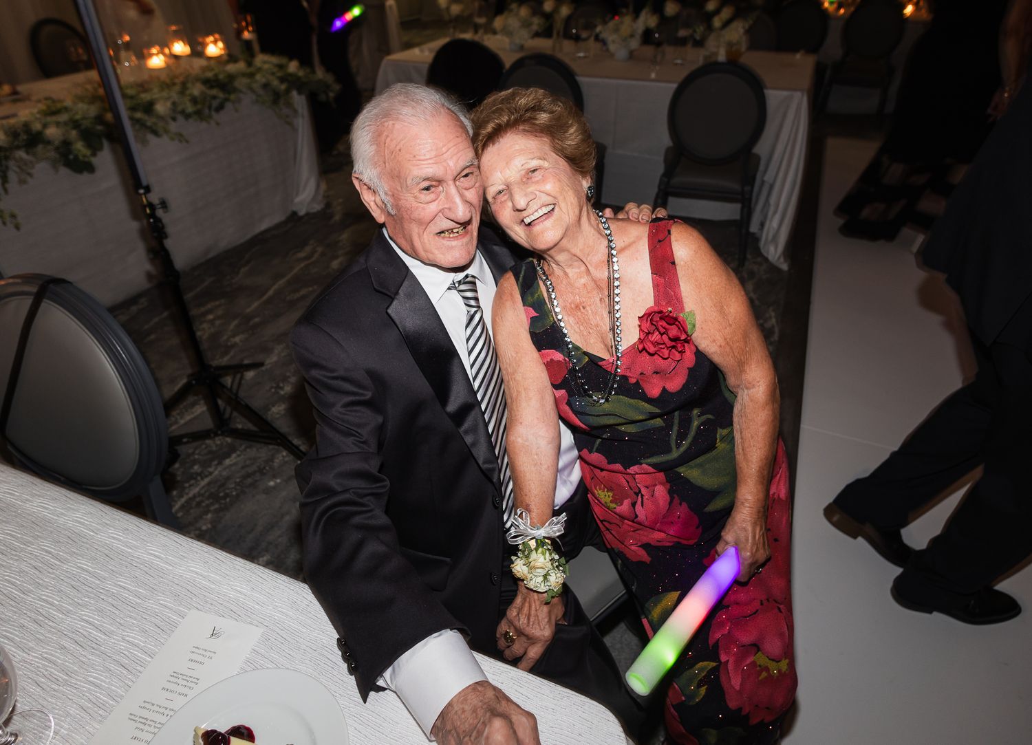 Elderly couple smiling and embracing at a wedding reception, woman holding a glow stick.