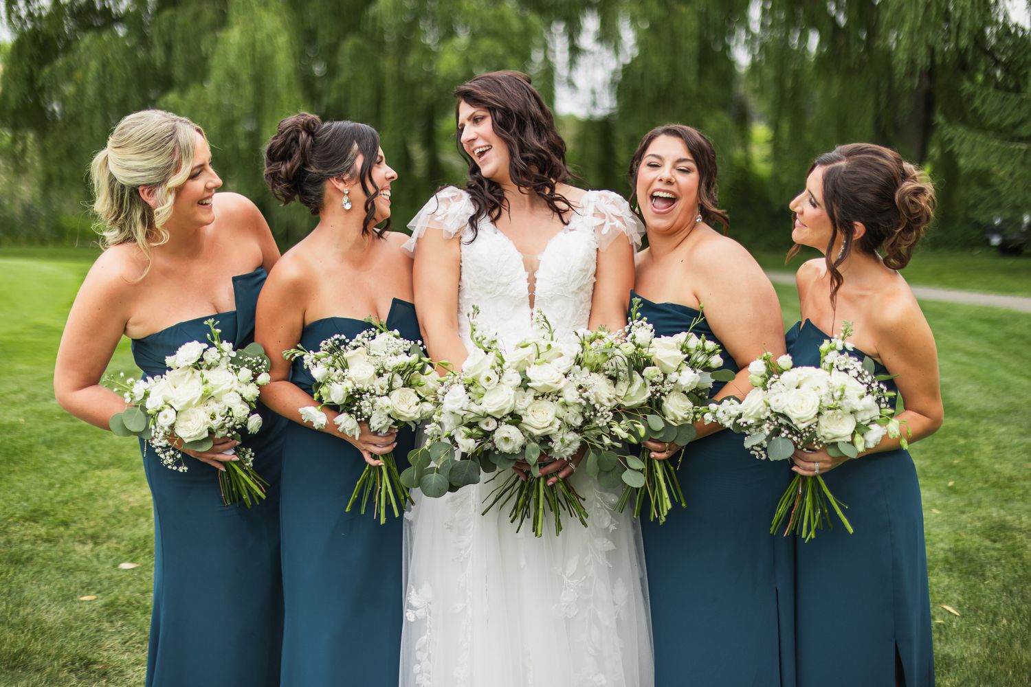 Bride in white lace gown laughs with bridesmaids in teal strapless dresses holding white floral bouquets.