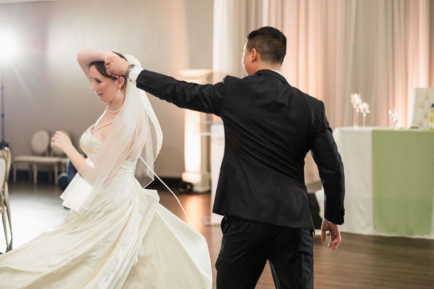 Bride and groom share their first dance at wedding reception, groom spinning bride in elegant white gown and veil.