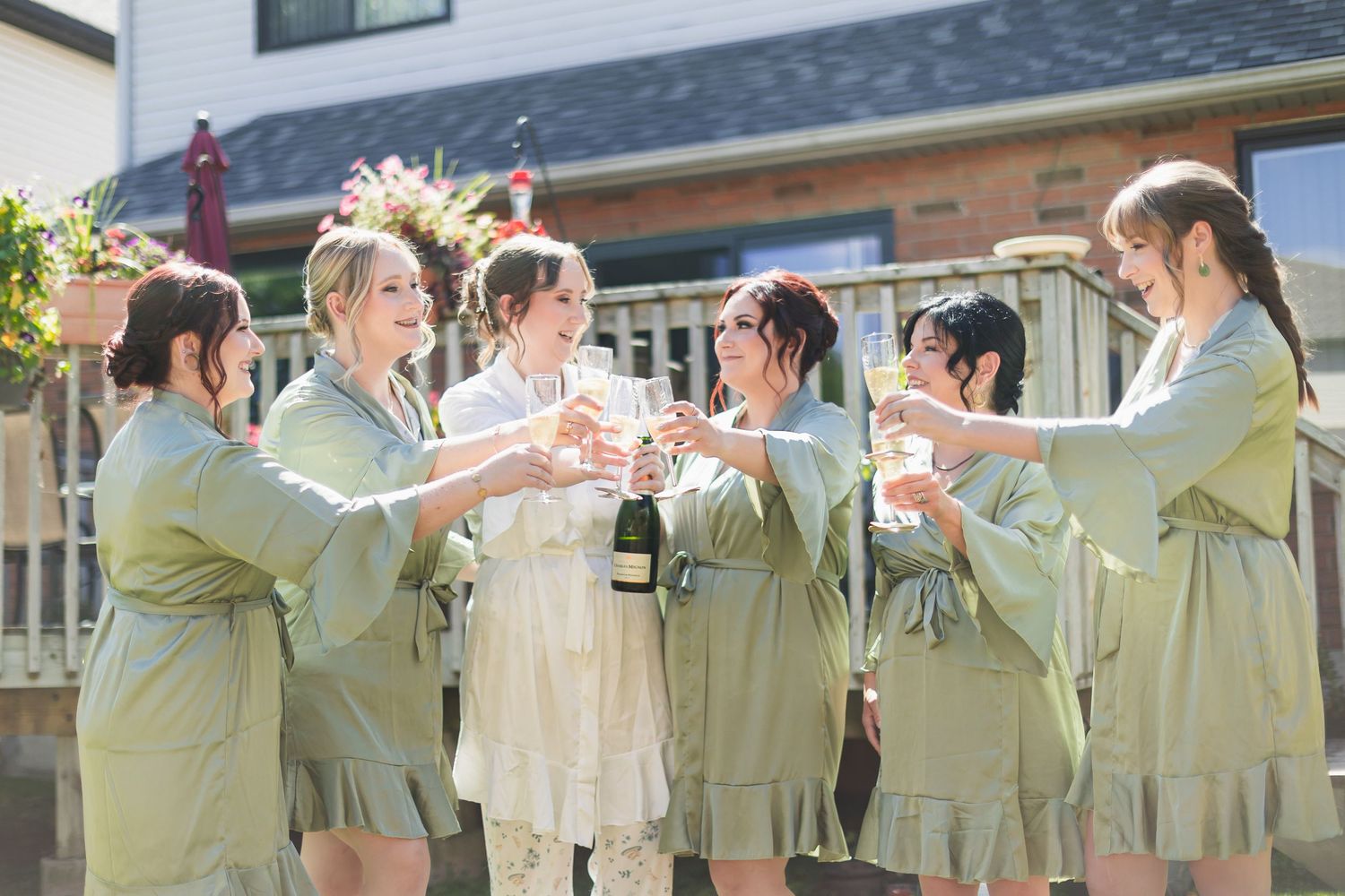 Bride and bridesmaids in sage green robes toast champagne together on sunny outdoor deck.