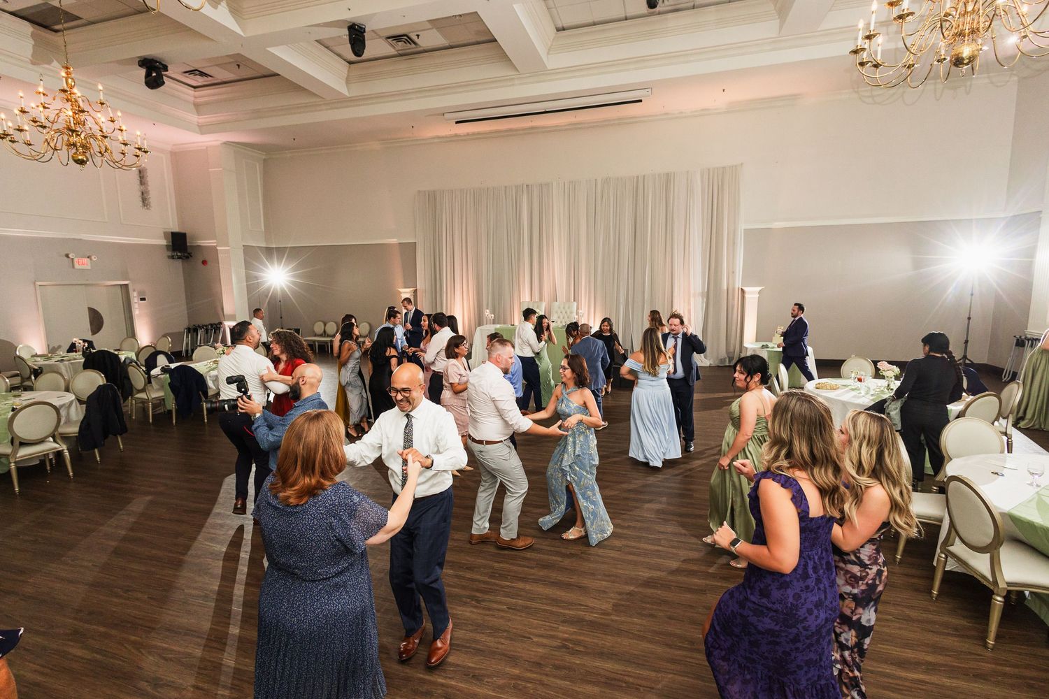 Guests dancing and socializing at an elegant wedding reception in a decorated ballroom with chandeliers.