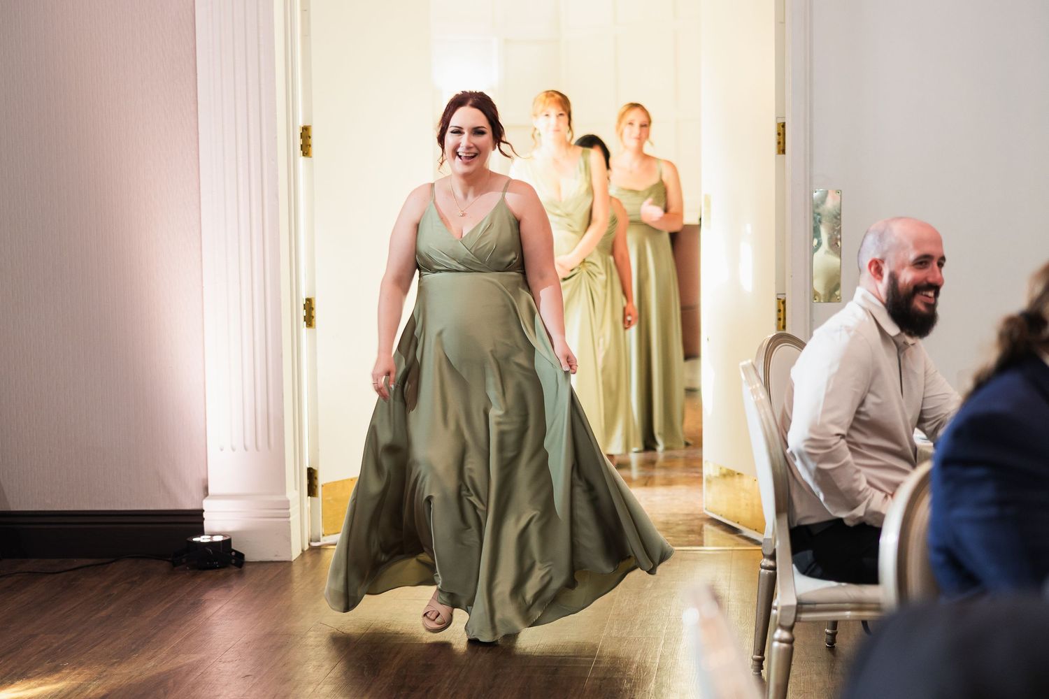 Bridesmaids in sage green gowns walking through elegant venue doorway during wedding processional.
