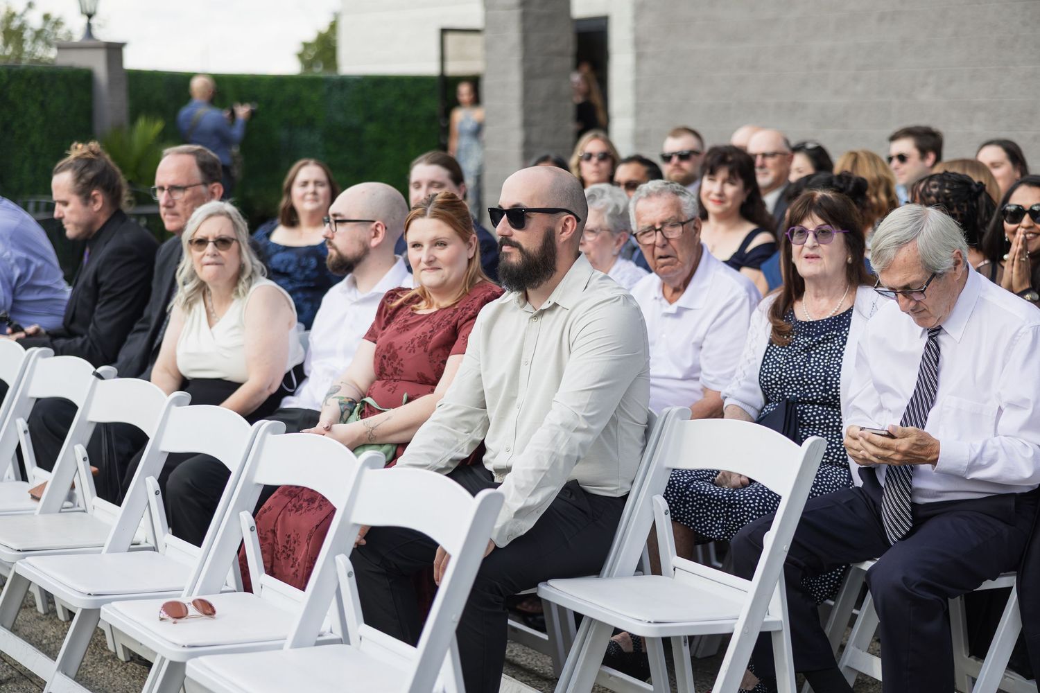 Wedding guests seated in white folding chairs during outdoor daytime ceremony.