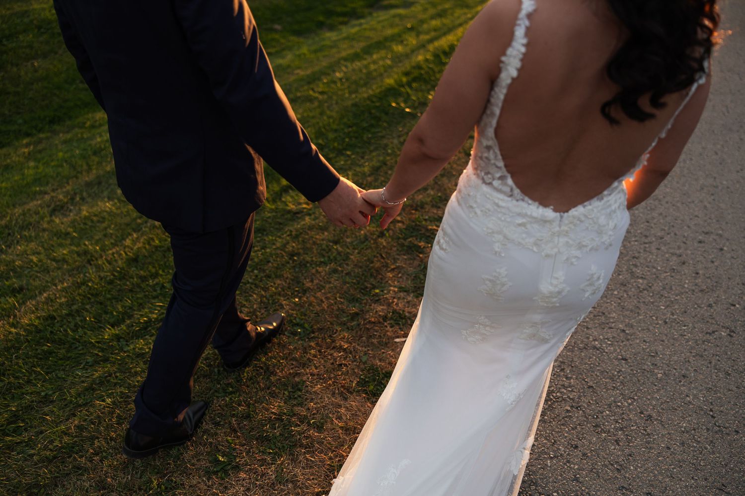 Bride and groom holding hands walking outdoors at golden hour, bride wearing elegant backless lace wedding gown.