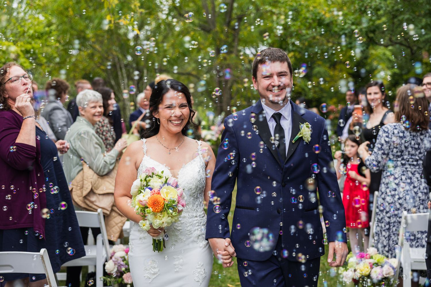 Newlywed couple walks down the aisle surrounded by bubbles as wedding guests celebrate their outdoor ceremony.