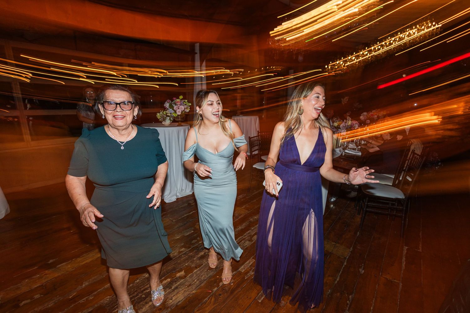 Three women dancing joyfully at a wedding reception in elegant dresses on a wooden dance floor.