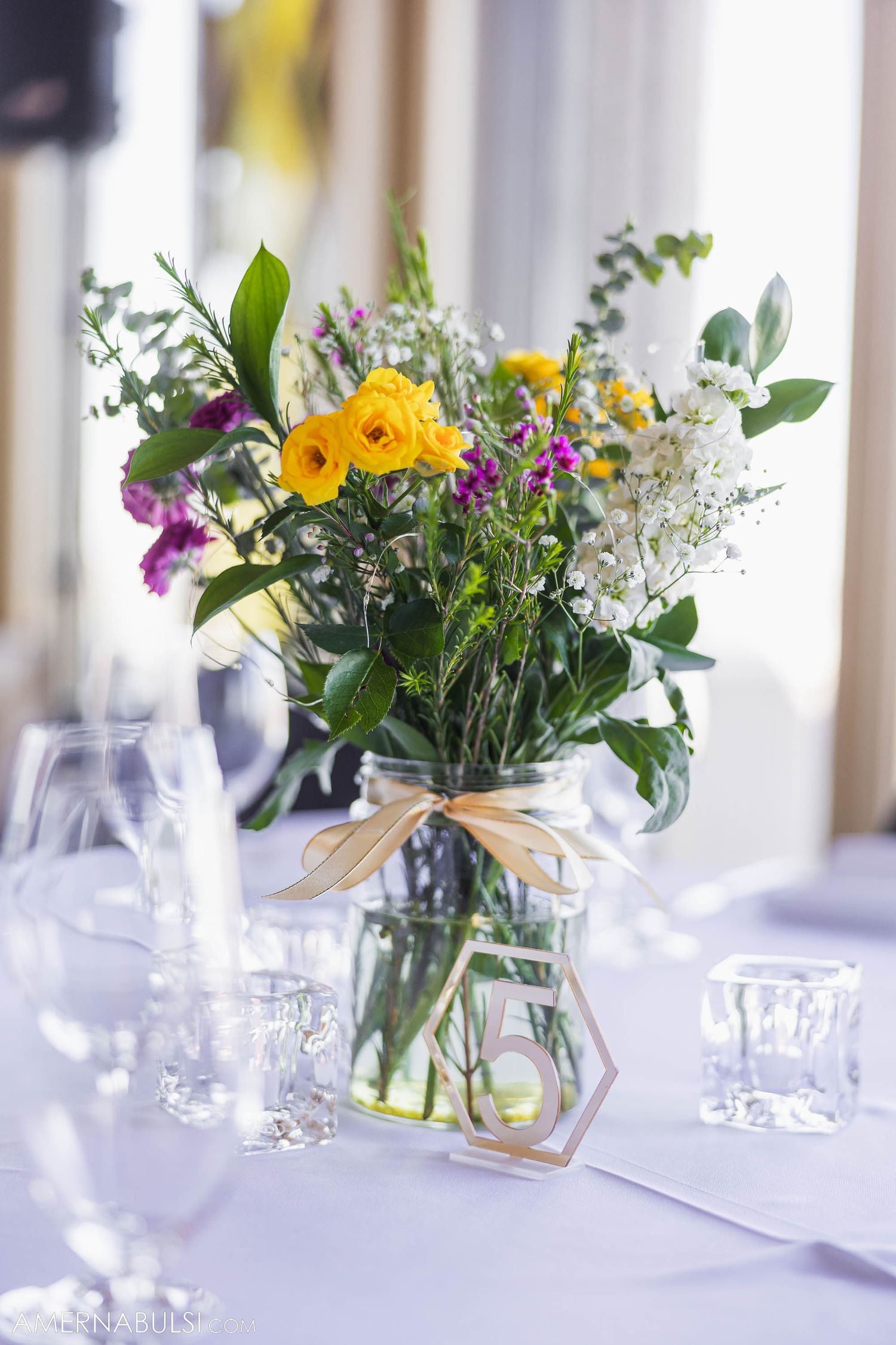 Colorful floral centerpiece with yellow roses and mixed wildflowers in a mason jar on a white tablecloth.