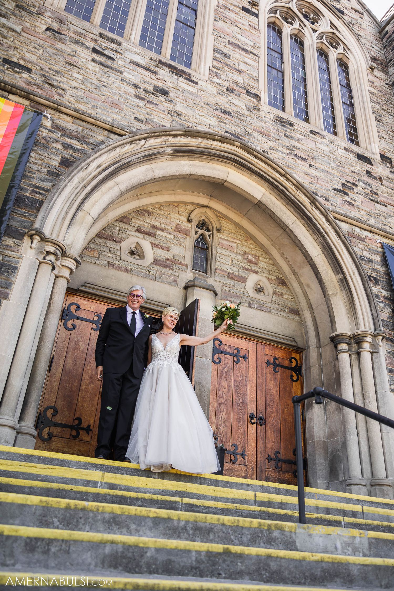A wedding couple poses under a stone archway entrance of a historic church.