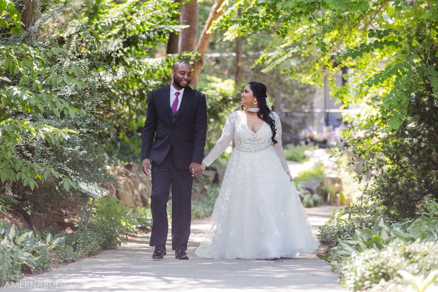 Couple walking hand in hand down tree-lined path in wedding attire.