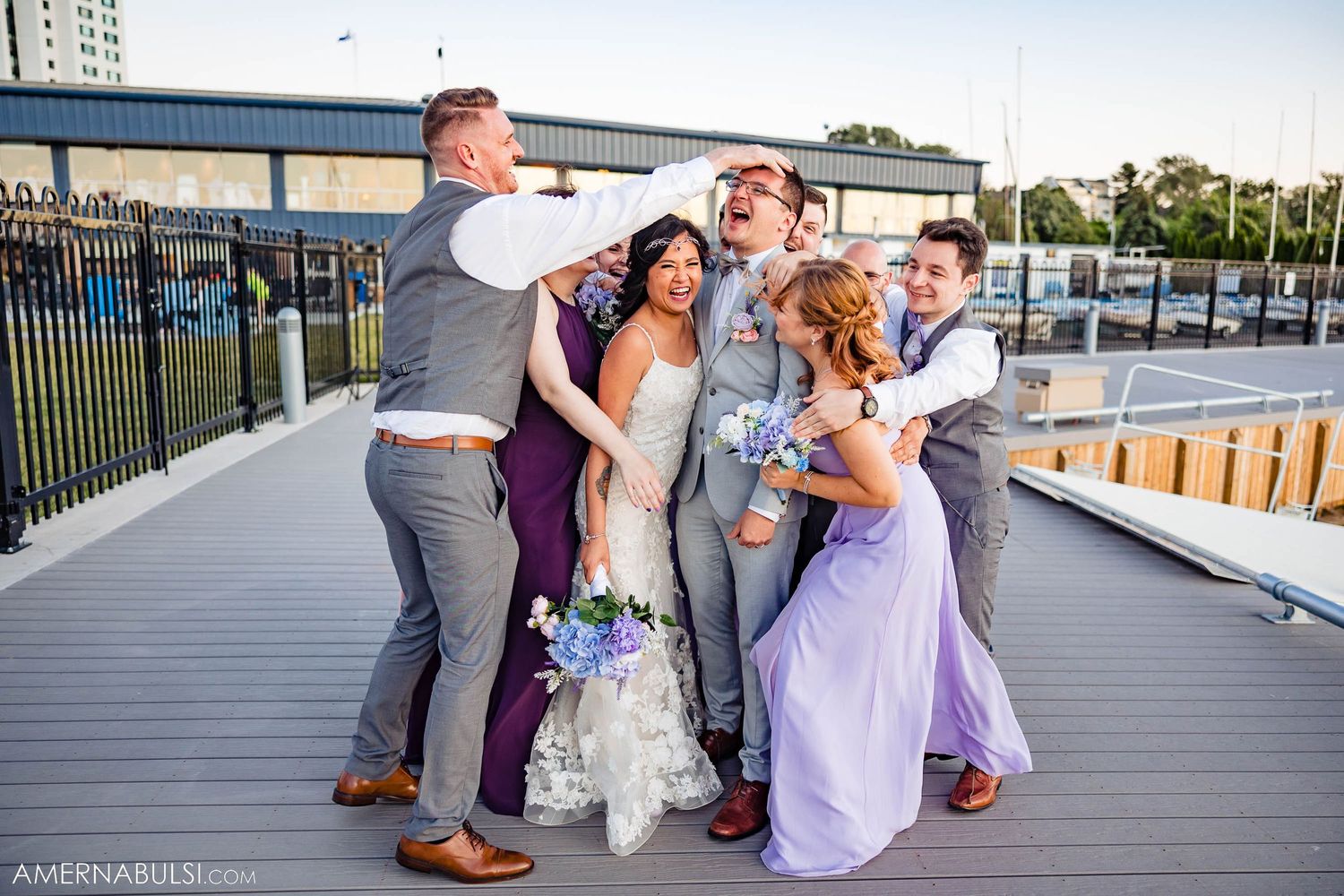 Joyful wedding party shares a group hug on marina deck with purple and grey color scheme.