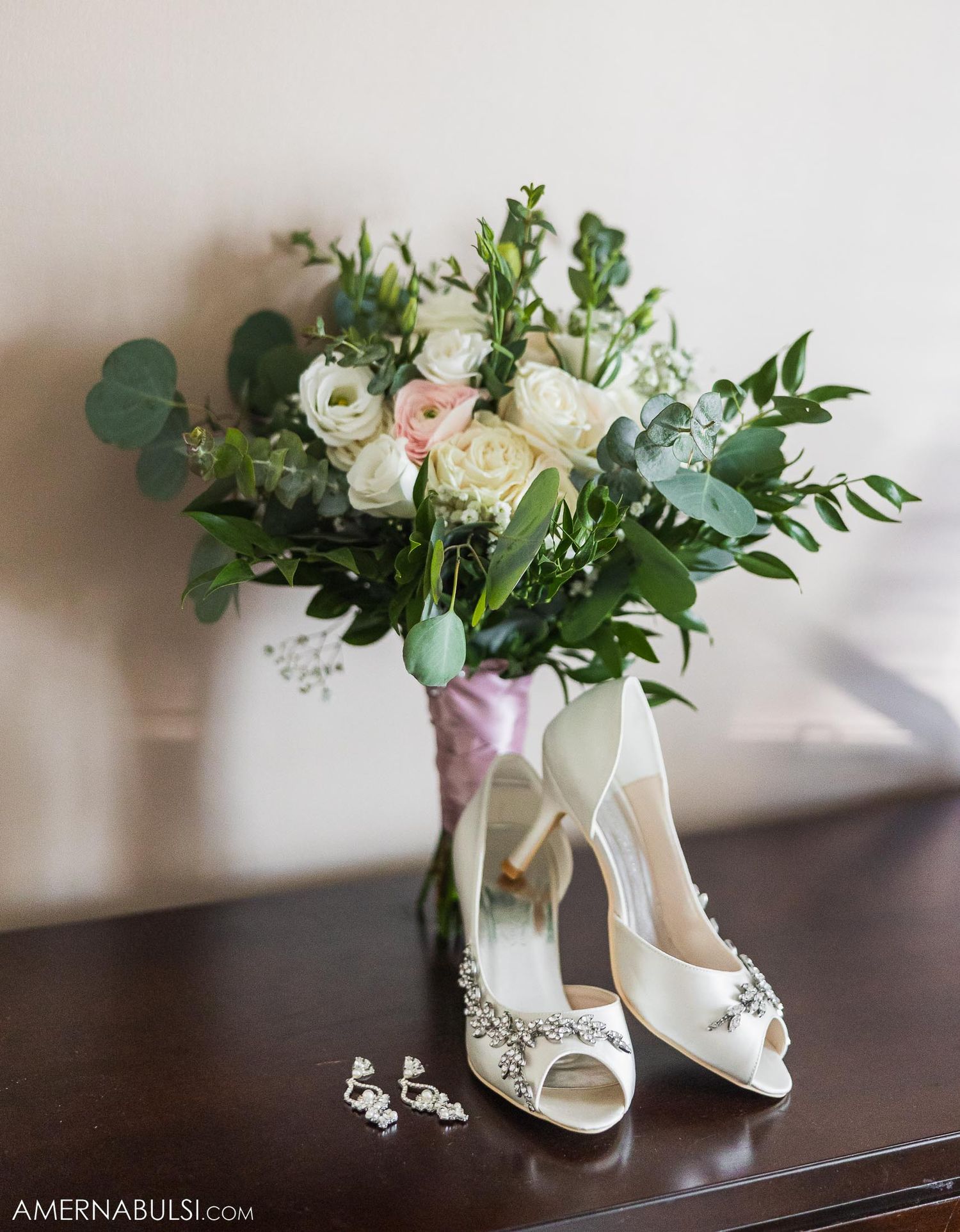 White bridal shoes and a delicate bouquet with roses and eucalyptus arranged on a wooden surface.