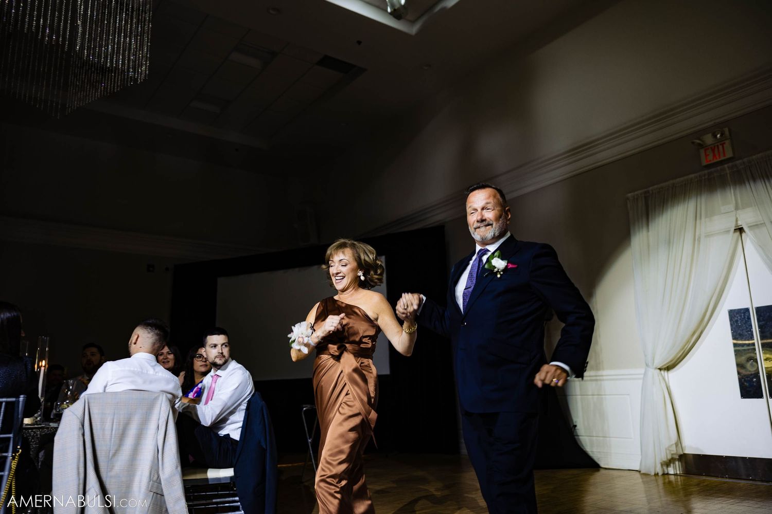 Attendees dance energetically at a wedding reception in an elegant venue with white curtains and dim lighting.