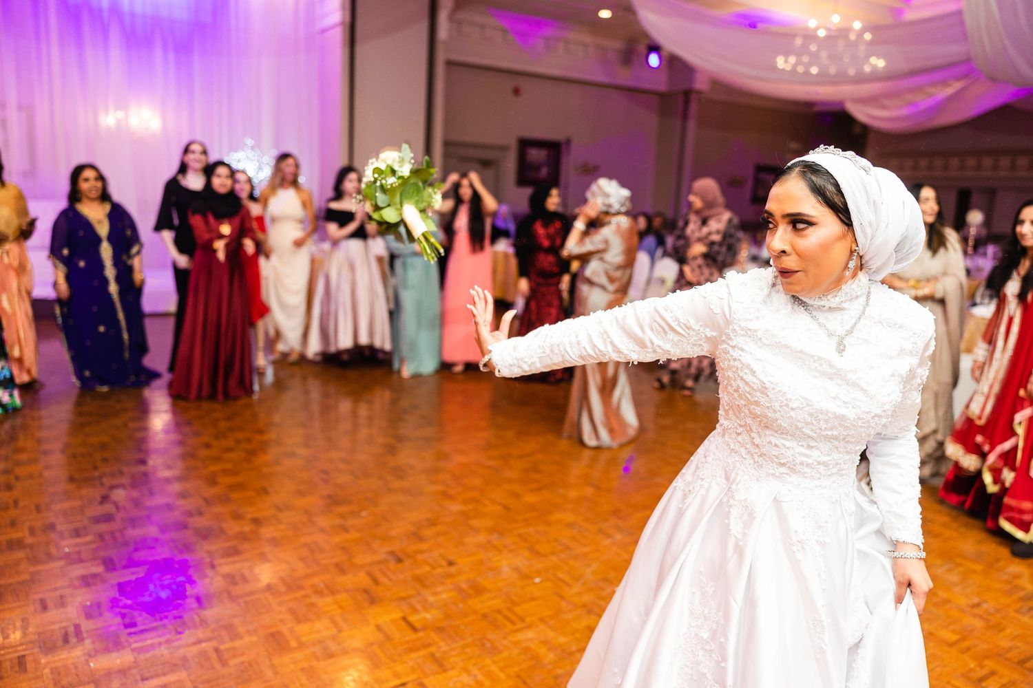 Person in white wedding dress dancing energetically at reception with dramatic lighting and wood floor.