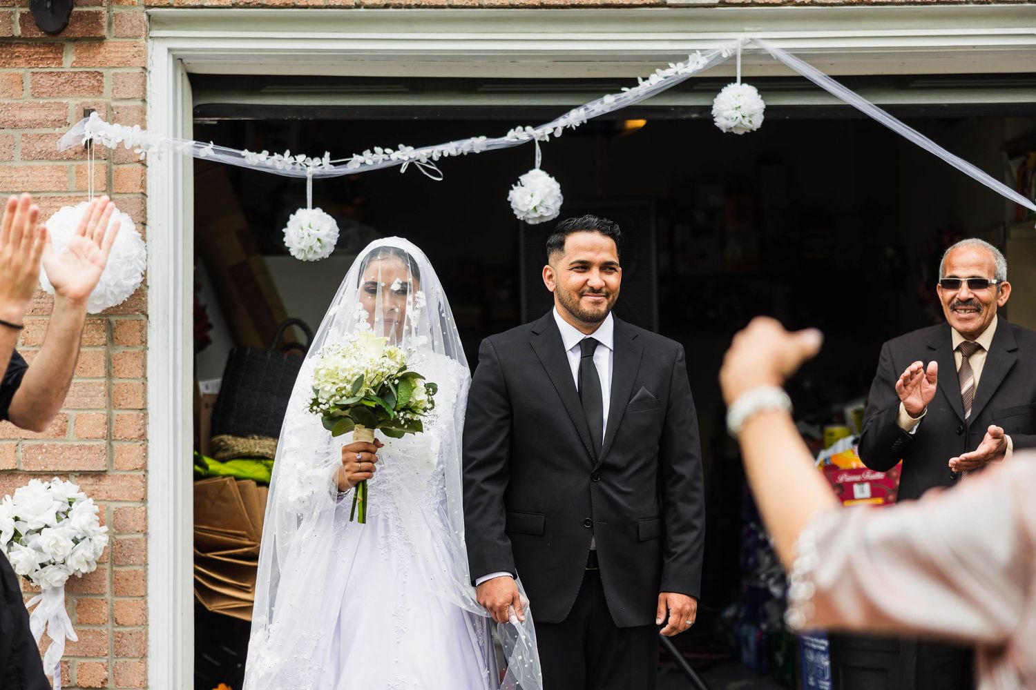 A couple exits their wedding venue under white lanterns and decorations while guests celebrate.