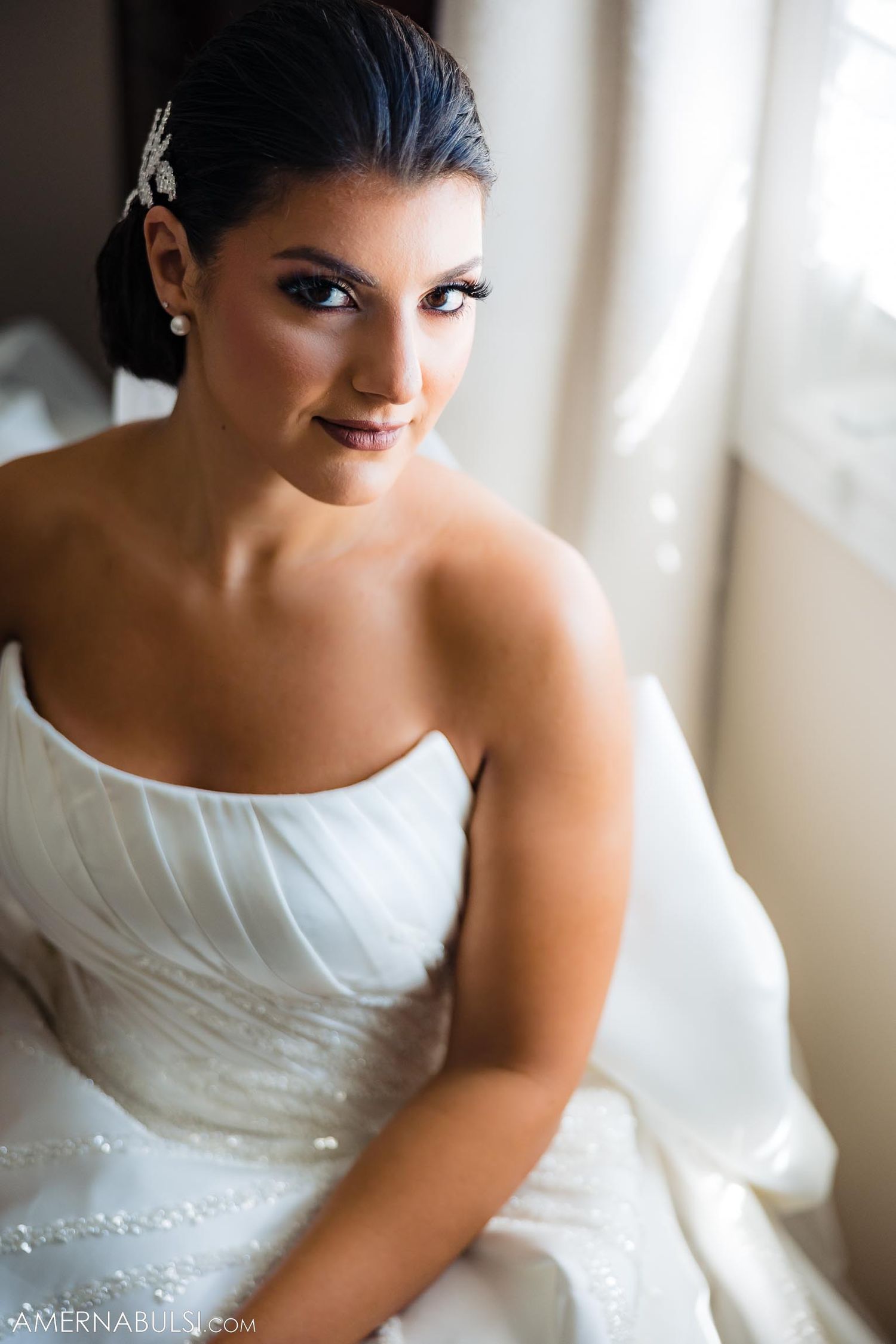 A bride in an elegant strapless gown poses by a window in natural light.